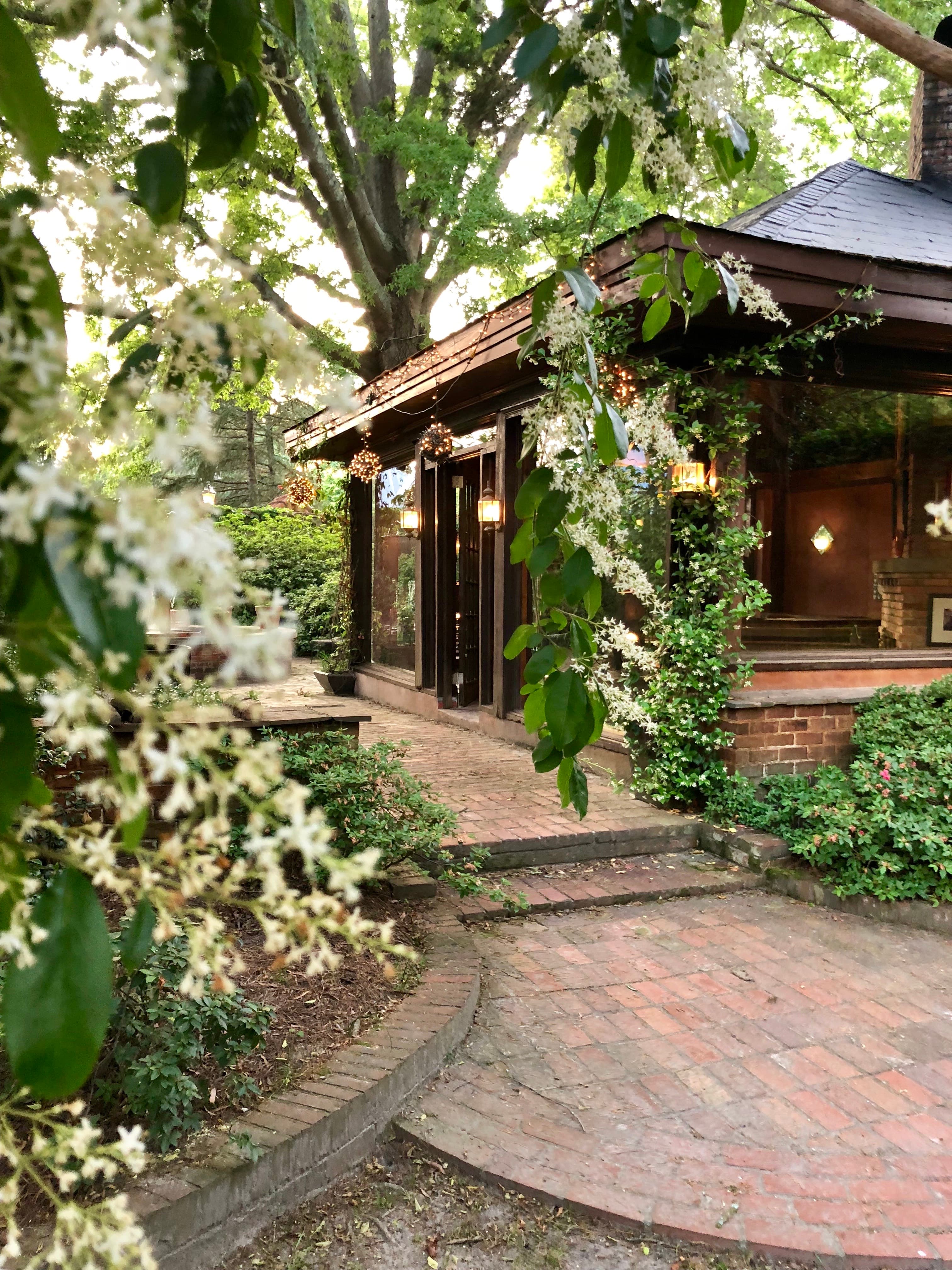 Brick path leading to a rustic wooden pavilion framed by white flowers and lush greenery.