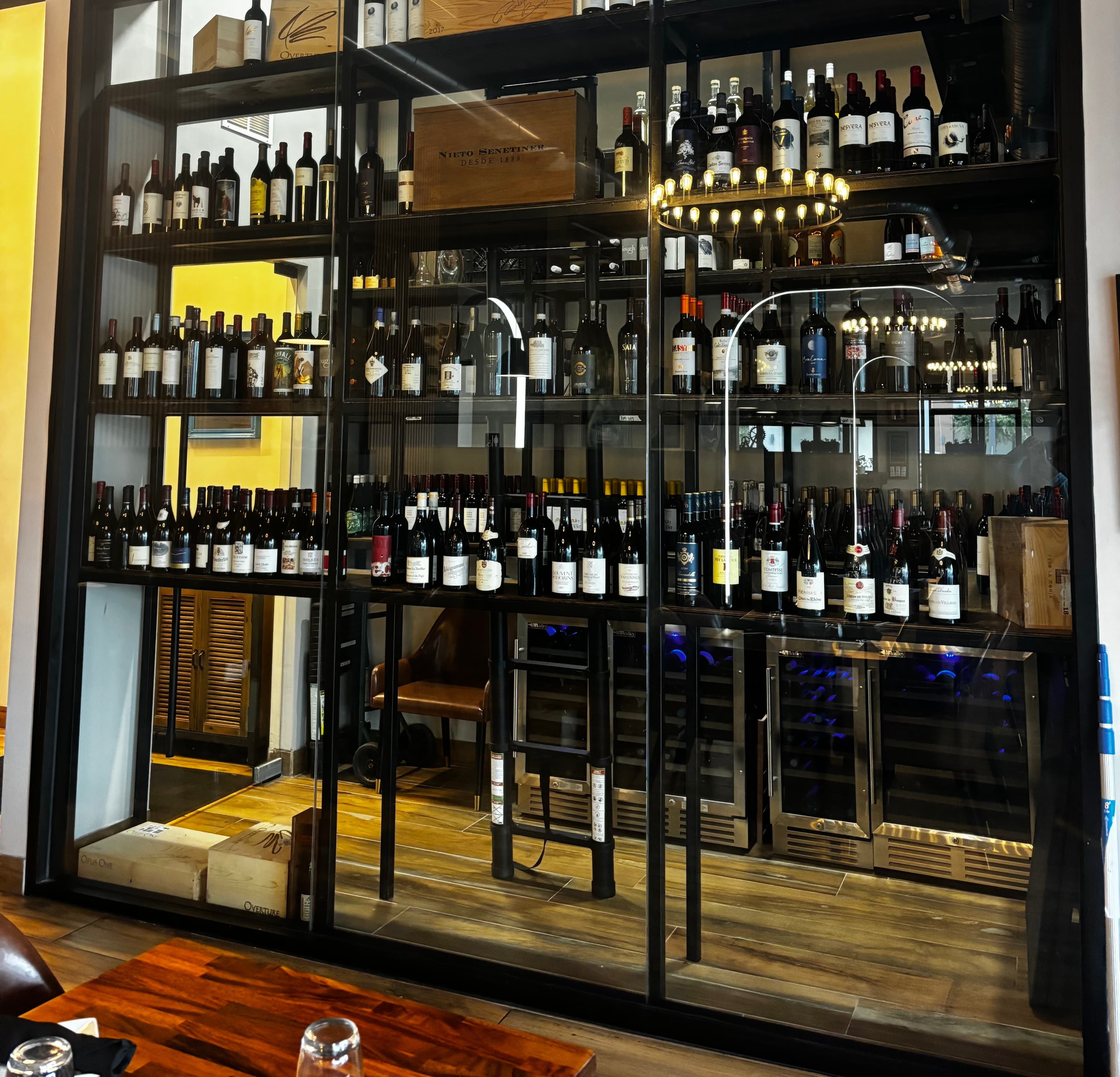 Large glass wine display case filled with bottles, wooden crates, and integrated refrigerated storage units.