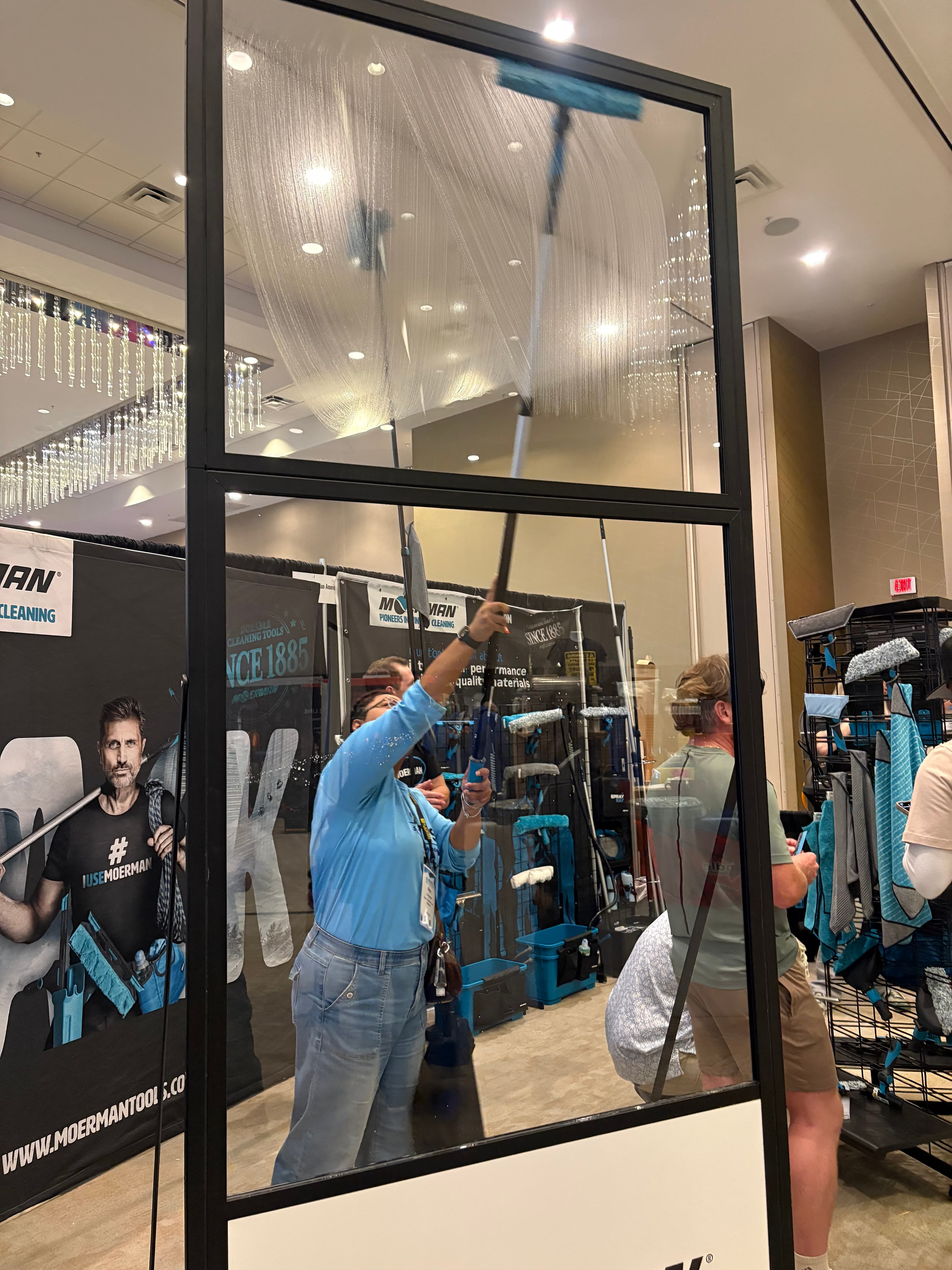 Person in blue shirt cleans a large soapy glass pane with a long-handled squeegee.