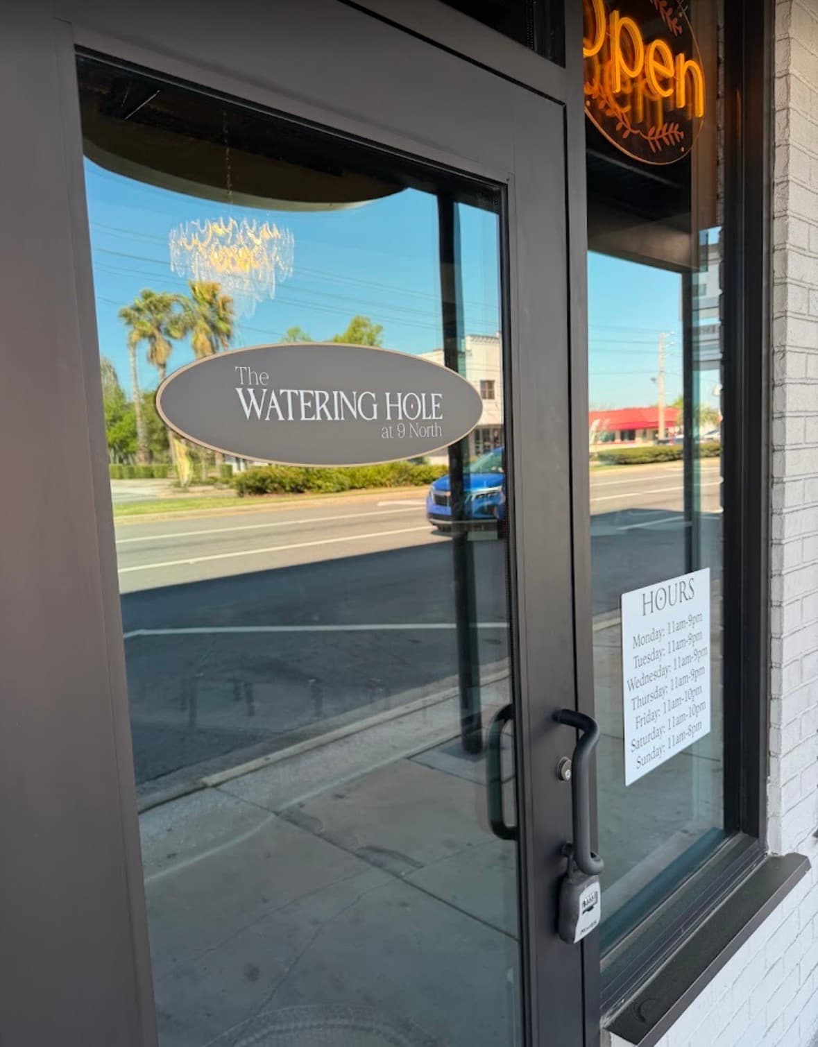 Glass door of The Watering Hole at 9 North showing business hours and street reflections.