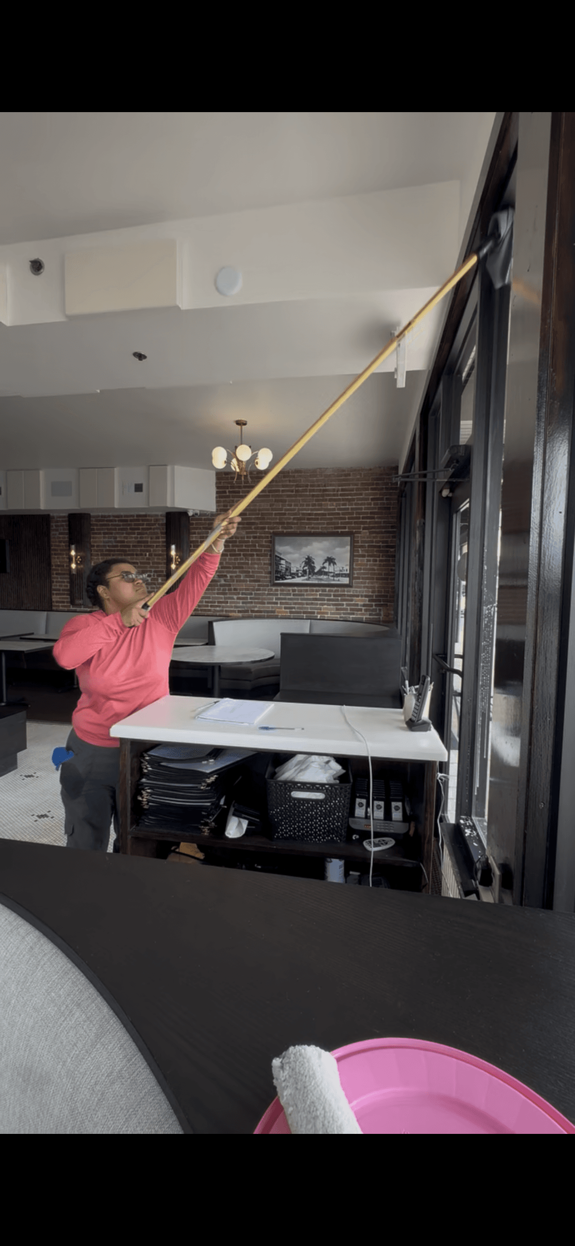 Woman in pink shirt uses a long yellow pole to clean high restaurant windows.