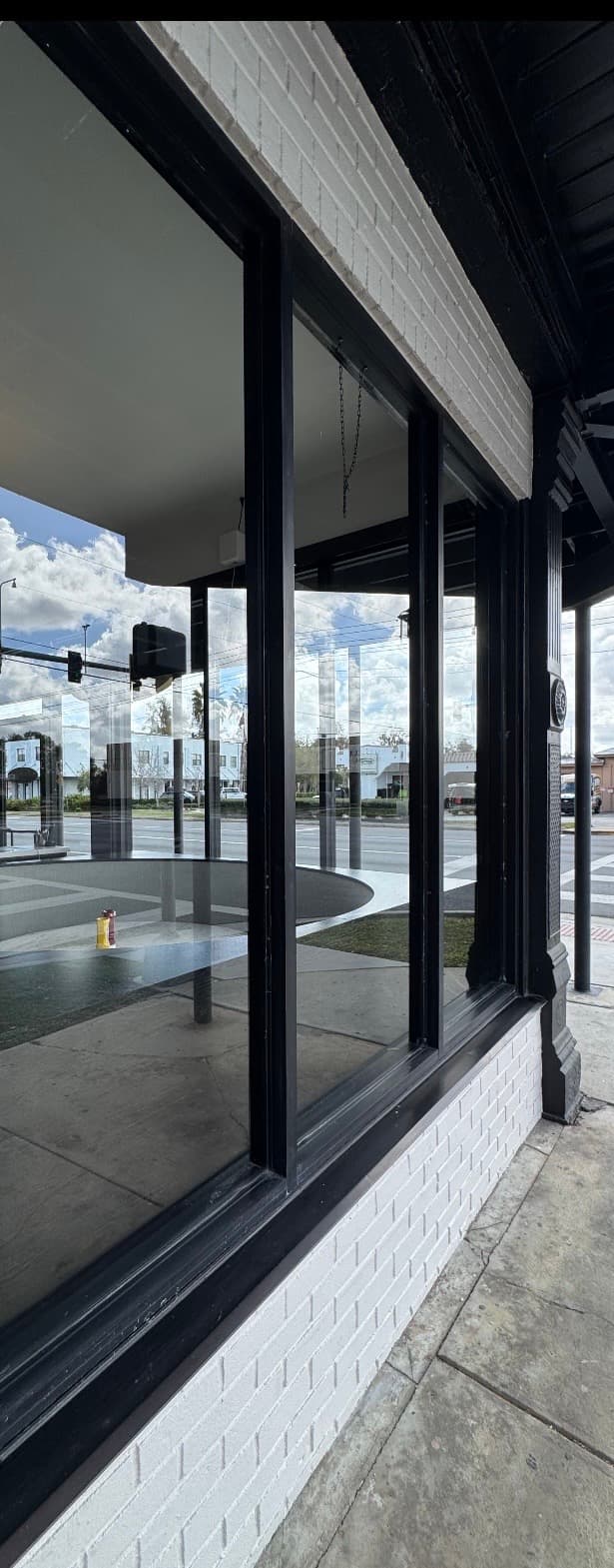 Storefront window with black frames reflecting a street and sky, set in white brick walls.
