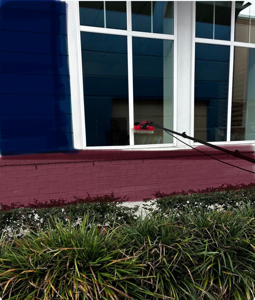 A long-handled red brush scrubbing a large window on a blue and brick building.